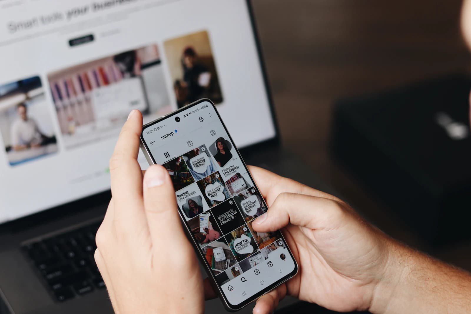 Person scrolling a social media feed on a smartphone in front of an open laptop