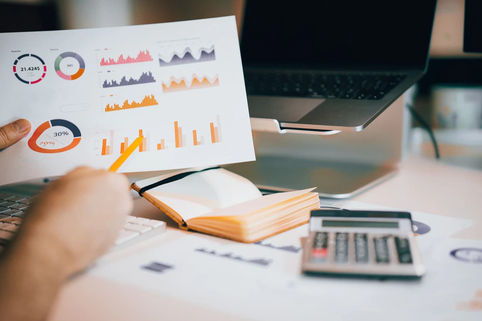 Person pointing a pencil at printed charts and graphs beside a laptop and notebook