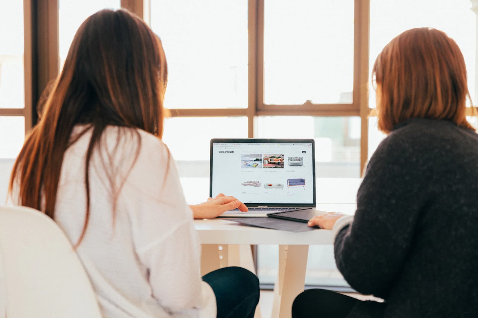 Two colleagues sitting at a table by a bright window, looking at a laptop together