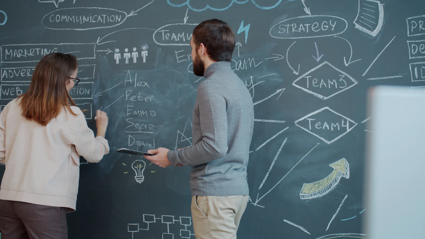 Two colleagues planning a marketing strategy on a large chalkboard wall covered in diagrams