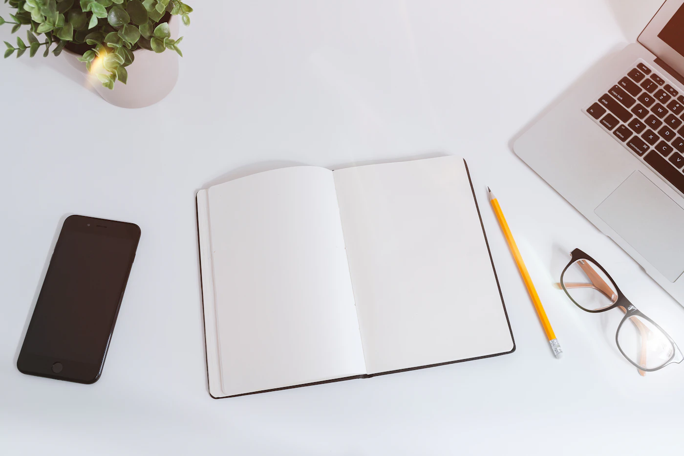 Open blank notebook with a pencil, glasses, phone and laptop on a white desk