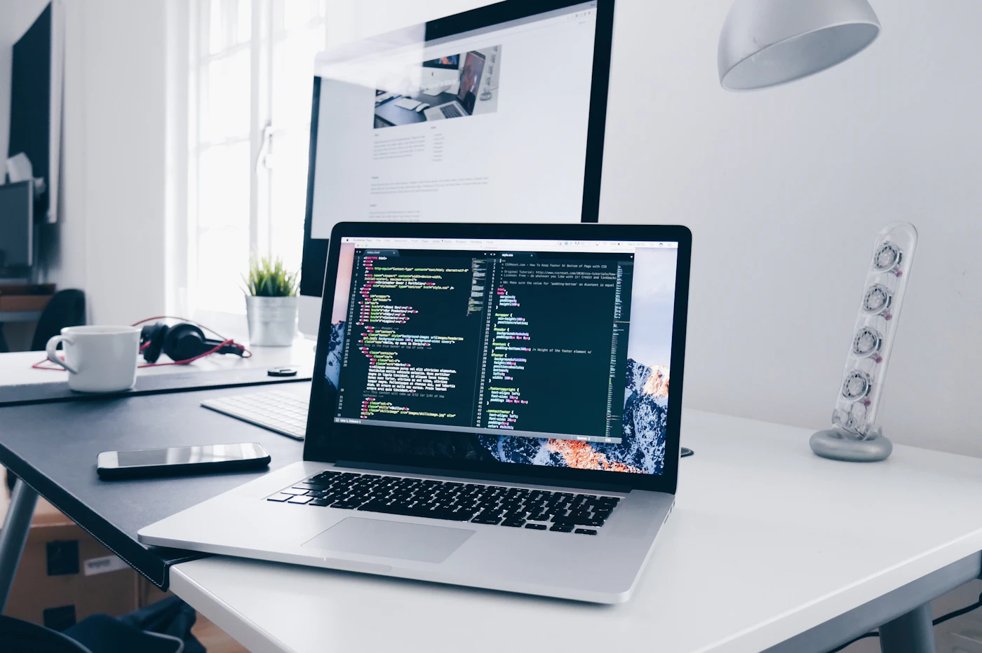 Laptop showing code next to a desktop monitor on a bright, tidy desk